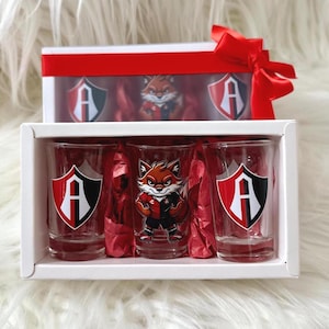May include: A gift set containing three shot glasses and a small figurine, each adorned with a fox mascot and the letter "A" on a shield. The items are presented in a white box with red detailing and a red bow.