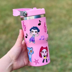May include: Pink insulated tumbler with a silver rim and a pink handle. The tumbler is decorated with musical notes and cartoon characters. The characters include a blue tiger, a girl with red hair, and a girl with dark hair.