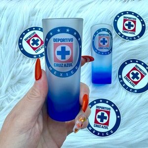 Puede incluir: Dos vasos de chupito con un dise&ntilde;o azul y blanco con el logotipo "DEPORTIVO CRUZ AZUL M&Eacute;XICO". El logotipo incluye una cruz azul con un contorno blanco. Los vasos de chupito est&aacute;n sobre una superficie blanca y peluda.