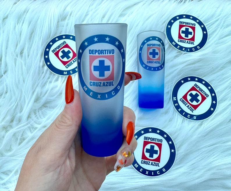 Puede incluir: Dos vasos de chupito con un dise&ntilde;o azul y blanco con el logotipo "DEPORTIVO CRUZ AZUL M&Eacute;XICO". El logotipo incluye una cruz azul con un contorno blanco. Los vasos de chupito est&aacute;n sobre una superficie blanca y peluda.