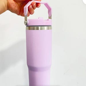 May include: A light purple insulated tumbler with a handle. The tumbler has a stainless steel band near the top and a matching light purple lid. The handle is also light purple and attached to the lid with silver metal hinges.
