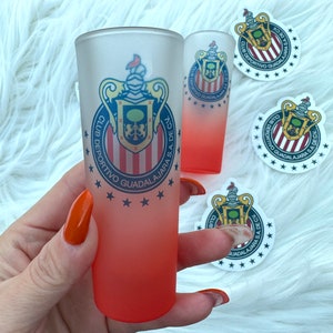Puede incluir: Dos vasos de chupito escarchados con un efecto degradado rojo. Los vasos presentan un logotipo blanco y rojo con el texto "CLUB DEPORTIVO GUADALAJARA S.A. DE C.V."