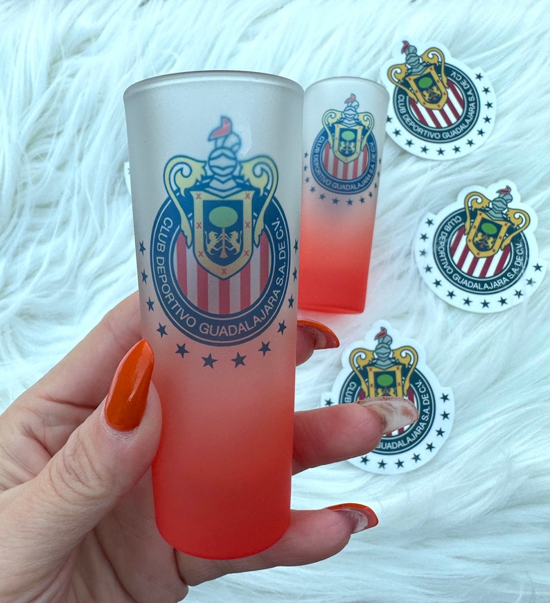 Puede incluir: Dos vasos de chupito escarchados con un efecto degradado rojo. Los vasos presentan un logotipo blanco y rojo con el texto "CLUB DEPORTIVO GUADALAJARA S.A. DE C.V."