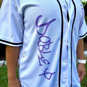 May include: White baseball jersey with black trim and a purple "Barbie" graphic on the front.