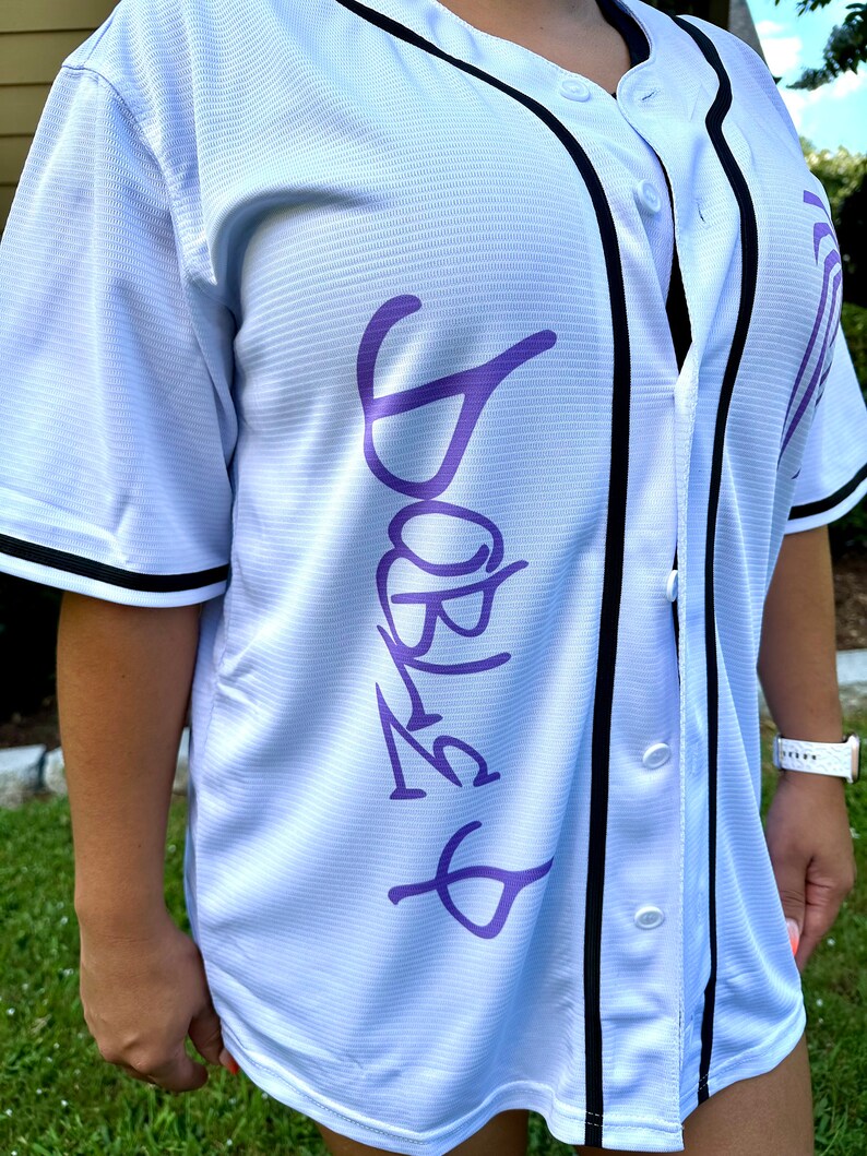 May include: White baseball jersey with black trim and a purple "Barbie" graphic on the front.