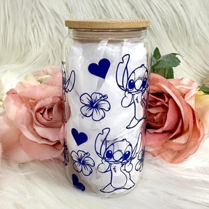 May include: A clear glass jar with a bamboo lid features a blue and white design of Stitch from Lilo & Stitch. The design includes hearts, hibiscus flowers, and Stitch in various poses.