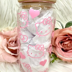 Puede incluir: Un tarro de vidrio transparente con un dise&ntilde;o de Hello Kitty rosa y una tapa de bamb&uacute;. El tarro est&aacute; lleno de papel de seda blanco y corazones rosas.
