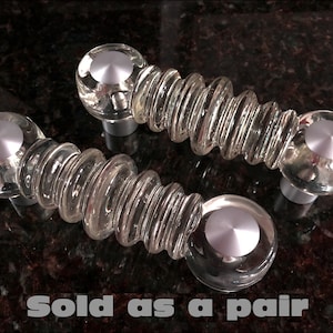 Pode incluir: Um par de botões de armário de vidro transparente com um centro de metal prateado. Os botões têm um design único e em camadas que se assemelha a uma espiral. O texto "Vendido em pares" é visível na imagem.