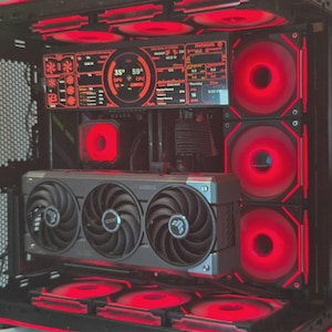 May include: A computer case interior featuring a horizontal graphics card with three black fans, multiple red LED cooling fans, and a rectangular digital display screen showing system performance metrics in red text against a black background.