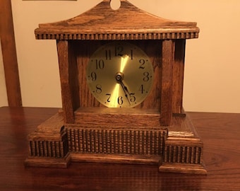 Può includere: Un orologio in legno con un quadrante dorato e numeri romani neri. L'orologio è in una cornice in legno decorativa con un piccolo tetto.