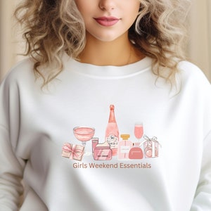 Könnte beinhalten: Weißer Pullover mit einer rosafarbenen Aquarell-Illustration einer Flasche Roséwein, einem Glas Rosé, einem Parfümflakon, einer Tube Tagescreme, einer Geschenkbox und einer kleinen Handtasche. Der Text "Girls Weekend Essentials" ist unter der Illustration gedruckt.