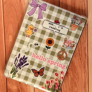 May include: Clear tablet case with a green and white gingham pattern, decorated with stickers including a purple bow, a duck holding a flower, a sunflower, and the text "hello spring".