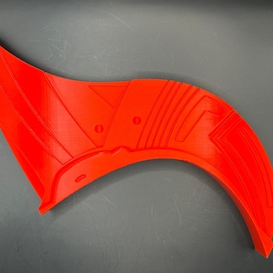 May include: A bright red, curved 3D-printed object with a wing-like shape. The object has a series of parallel lines and circular indentations. The design features a series of geometric shapes and a curved edge.