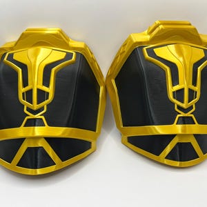 May include: A pair of black and gold plastic shoulder pads with a geometric design. The shoulder pads are made of a smooth, shiny plastic.