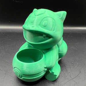 Peut inclure: Un pot de fleurs imprimé en 3D de couleur verte en forme de personnage de dessin animé. Le pot a une ouverture ronde en haut pour la plantation.