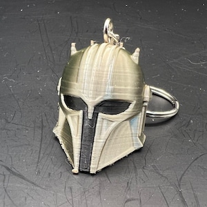 May include: A 3D printed Mandalorian helmet keyring in a light grey colour with black details. The keyring has a silver metal ring.