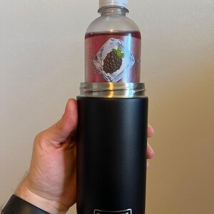 Puede incluir: Una botella YETI Rambler negra de 12 oz con una tapa negra y una botella de pl&aacute;stico transparente con un l&iacute;quido rojo en su interior. La botella de pl&aacute;stico tiene una etiqueta con un gr&aacute;fico de mora y un cubito de hielo.