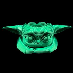 May include: A green 3D printed planter shaped like a character from the Star Wars franchise. The planter has a large head with big eyes and a small mouth.
