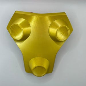 May include: A gold-colored plastic object with three round, indented knobs. The object is shaped like a rounded triangle.