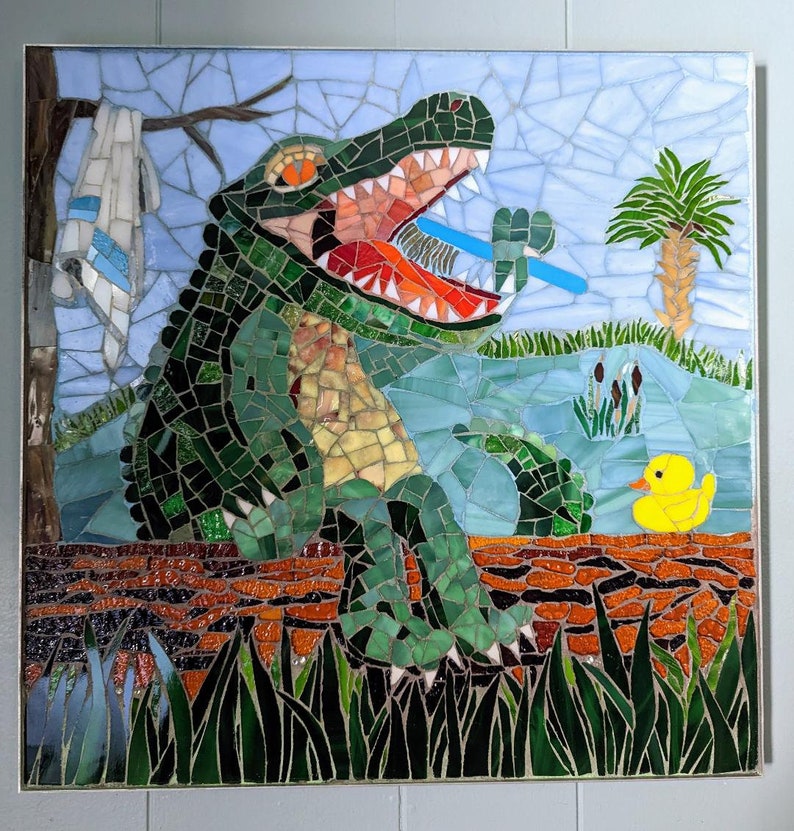 May include: A mosaic art piece depicting a green alligator with its mouth open, holding a blue and green object in its teeth. The alligator is sitting on a brown log with green grass in the foreground. A yellow rubber duck is floating in the water behind the alligator.