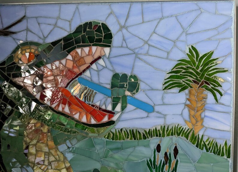 May include: A mosaic artwork depicting a green alligator with its mouth open, holding a blue toothbrush. The alligator is in a swamp setting with green grass and a palm tree.