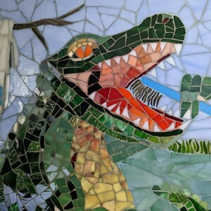 May include: A mosaic artwork depicting a green alligator with an open mouth, revealing sharp teeth. The alligator is biting down on a blue object, possibly a toothbrush. The background is a light blue with a white cloth hanging from a branch.