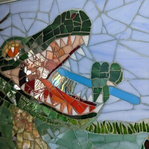 May include: A mosaic artwork depicting a green alligator with an open mouth, revealing sharp teeth. The alligator is biting down on a blue toothbrush. The background is a light blue mosaic with green foliage.