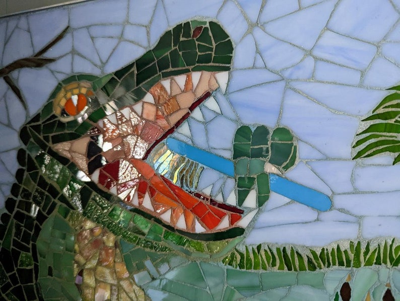 May include: A mosaic artwork depicting a green alligator with an open mouth, revealing sharp teeth. The alligator is biting down on a blue toothbrush. The background is a light blue mosaic with green foliage.