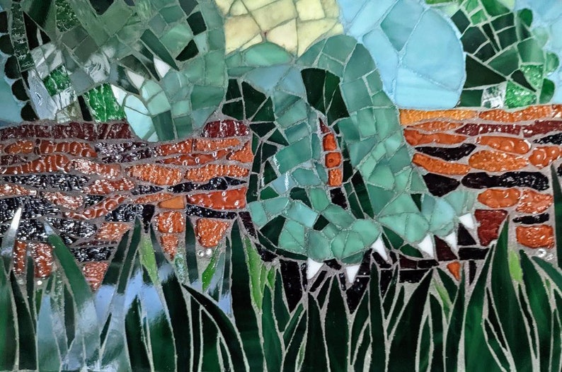 May include: A close-up of a mosaic artwork depicting a green leafy plant with a brown brick wall in the background. The mosaic is made of small, colorful glass tiles.