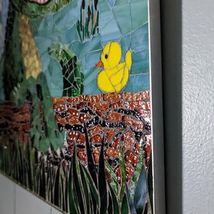 May include: A mosaic artwork featuring a yellow rubber duck, green grass, and a brown brick wall. The artwork is framed with a silver metal frame.