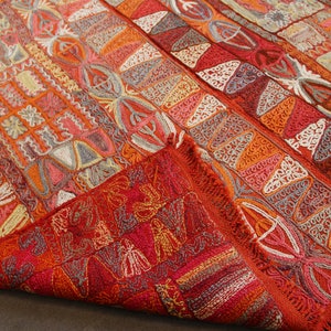 Pu&ograve; includere: Un tappeto colorato tessuto a mano con intricati motivi geometrici in tonalit&agrave; di rosso, arancione, bianco e grigio. Il tappeto ha un bordo con frange ed &egrave; piegato a met&agrave;.
