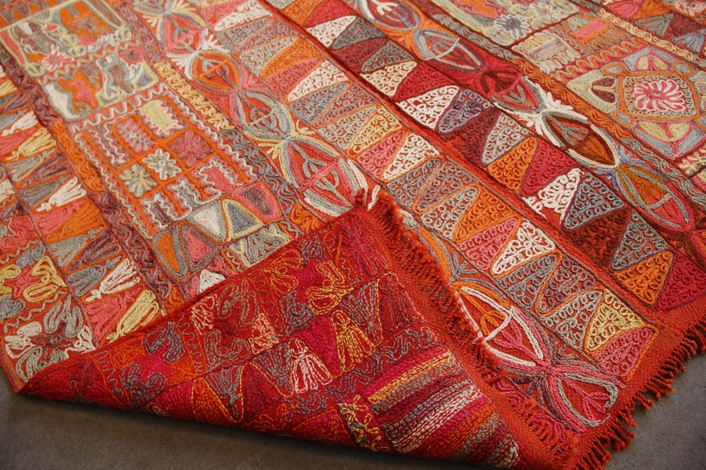Pu&ograve; includere: Un tappeto colorato tessuto a mano con intricati motivi geometrici in tonalit&agrave; di rosso, arancione, bianco e grigio. Il tappeto ha un bordo con frange ed &egrave; piegato a met&agrave;.