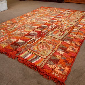 Pu&ograve; includere: Un grande tappeto con un motivo intricato con una miscela di colori arancio, rosso, marrone e bianco. Il tappeto ha un bordo a frangia e un design geometrico.
