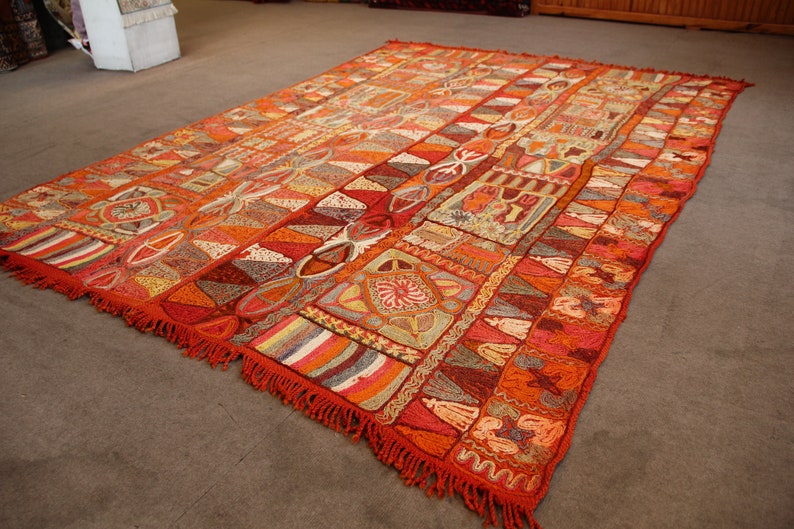 Pu&ograve; includere: Un grande tappeto con un motivo intricato con una miscela di colori arancio, rosso, marrone e bianco. Il tappeto ha un bordo a frangia e un design geometrico.