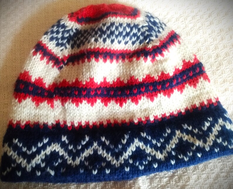 May include: A knitted beanie hat with a geometric pattern in red, white, and navy blue. The hat features a ribbed brim and a repeating zigzag design. The top of the hat is red, with white and navy blue accents.