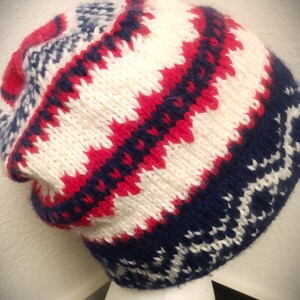 May include: A knitted beanie hat with a geometric pattern in red, white, and navy blue. The hat features a folded brim and a textured knit design, perfect for cold weather. The hat is displayed on a white mannequin head.
