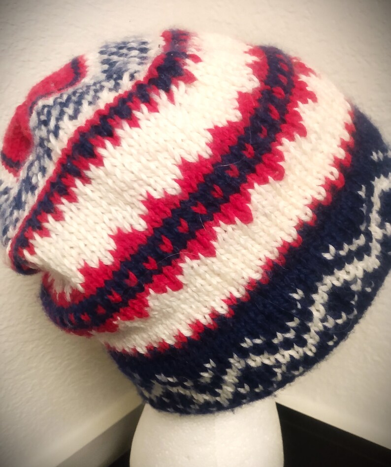 May include: A knitted beanie hat with a geometric pattern in red, white, and navy blue. The hat features a folded brim and a textured knit design, perfect for cold weather. The hat is displayed on a white mannequin head.