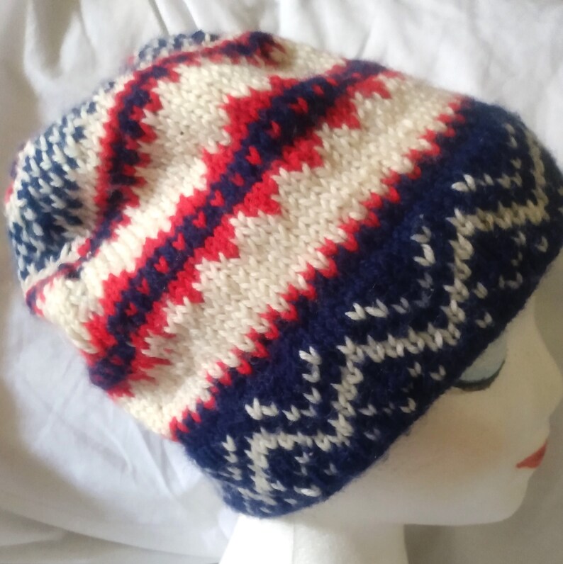 May include: A knitted beanie hat with a geometric pattern in red, white, and navy blue. The hat features a zig-zag design across the crown and a solid navy blue band at the bottom. The hat is made of a soft, warm material.