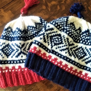 May include: Two knitted winter hats with a geometric pattern. The hats are white, navy blue, and red. One hat has a red tassel, and the other has a navy blue pom-pom on top. The hats are on a wooden surface.