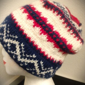 May include: A knitted beanie hat in a fair isle pattern. The hat features a color scheme of white, navy blue, and red. The hat is displayed on a mannequin head. The hat has a folded brim and a slouchy fit.