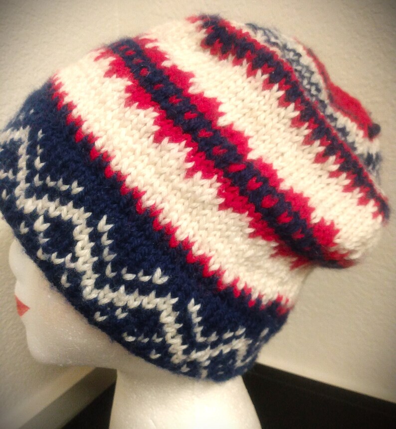 May include: A knitted beanie hat in a fair isle pattern. The hat features a color scheme of white, navy blue, and red. The hat is displayed on a mannequin head. The hat has a folded brim and a slouchy fit.