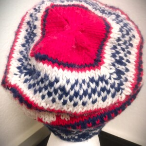 May include: A knitted hat with a red top and a pattern of white, navy blue, and red. The hat is made of a soft, textured yarn and has a rounded shape. The hat is designed to keep the head warm and is suitable for cold weather.