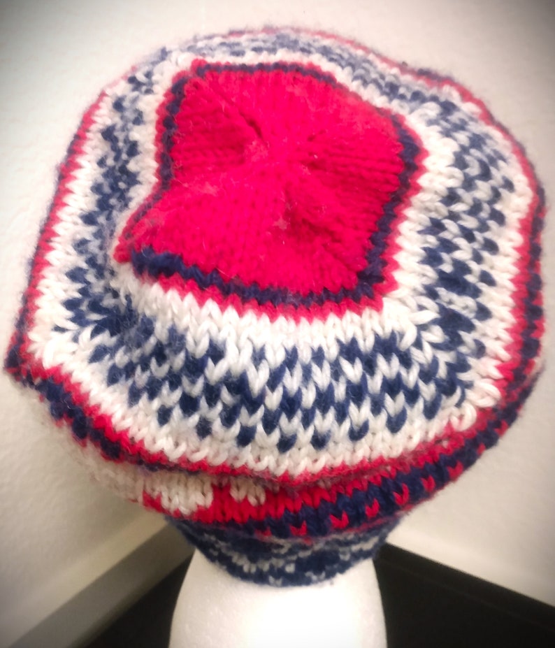 May include: A knitted hat with a red top and a pattern of white, navy blue, and red. The hat is made of a soft, textured yarn and has a rounded shape. The hat is designed to keep the head warm and is suitable for cold weather.