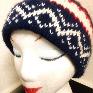 May include: A knitted beanie hat in navy blue, white, and red. The hat features a geometric pattern in white against the navy blue base, with red and white stripes near the top. The hat is displayed on a mannequin head.