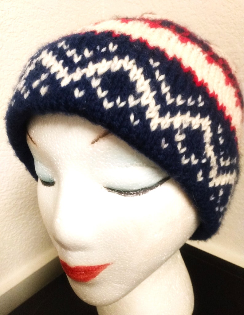 May include: A knitted beanie hat in navy blue, white, and red. The hat features a geometric pattern in white against the navy blue base, with red and white stripes near the top. The hat is displayed on a mannequin head.