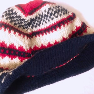 May include: A knitted beanie hat with a geometric pattern in red, navy blue, and cream colors. The hat features a folded navy blue brim and a textured knit design. This winter accessory is suitable for cold weather.