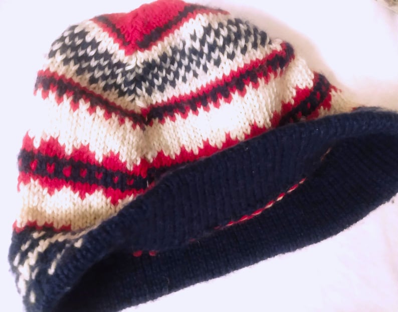 May include: A knitted beanie hat with a geometric pattern in red, navy blue, and cream colors. The hat features a folded navy blue brim and a textured knit design. This winter accessory is suitable for cold weather.