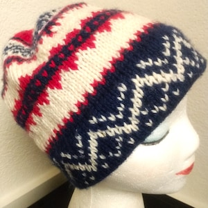 May include: A knitted beanie hat with a geometric pattern. The hat features alternating bands of white, navy blue, and red. The navy blue section has a white snowflake-like design. The hat is displayed on a white mannequin head.