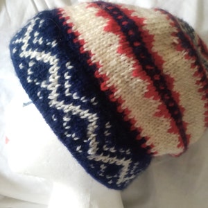 May include: A knitted beanie hat with a geometric pattern. The hat features a navy blue base with white snowflake-like designs, and a cream and red striped pattern on the crown. The hat is displayed on a white mannequin head.