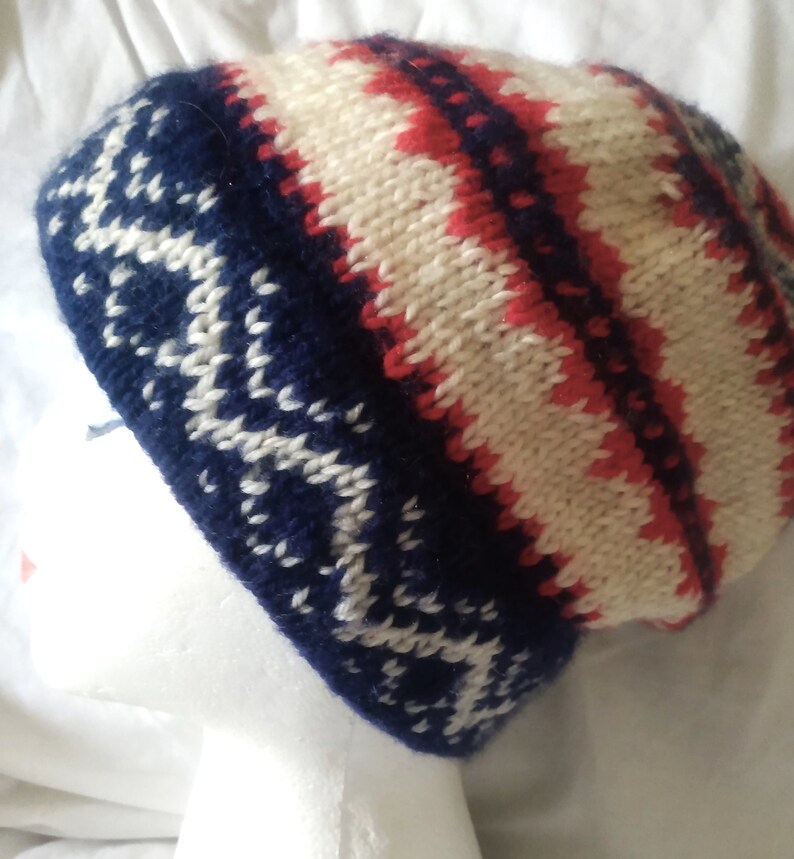 May include: A knitted beanie hat with a geometric pattern. The hat features a navy blue base with white snowflake-like designs, and a cream and red striped pattern on the crown. The hat is displayed on a white mannequin head.